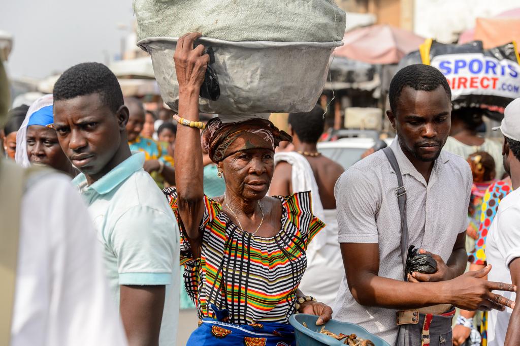 A woman carries a large sack on her head while walking through a busy market, flanked by two men